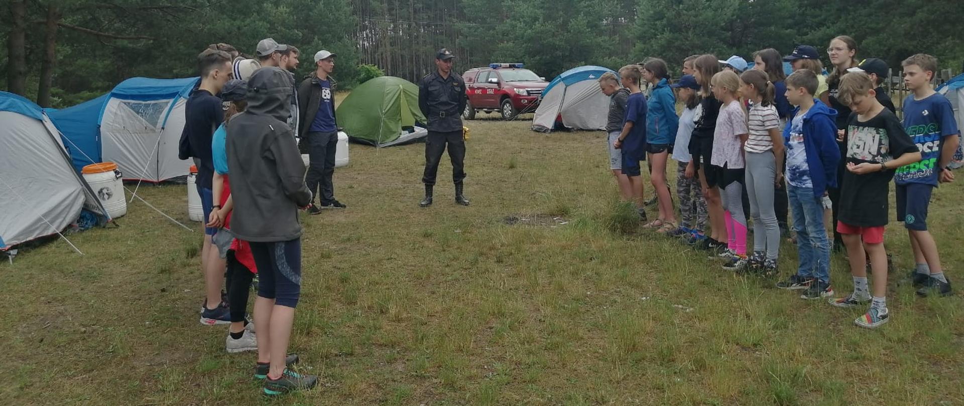 Zdjęcie obrazuje obóz harcerski usytuowany w lesie. Na pierwszym palnie widać grupę dzieci, którzy słuchają strażaka w umundurowaniu kadry dowódczo-sztabowej. Na drugim planie widnieją namioty oraz samochód operacyjny Państwowej Straży Pożarnej.
