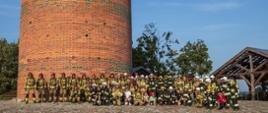 Zdjęcie grupowe strażaków w umundurowaniu specjalnym stojących u podstawy wieży wykonanej z czerwonej cegły.
