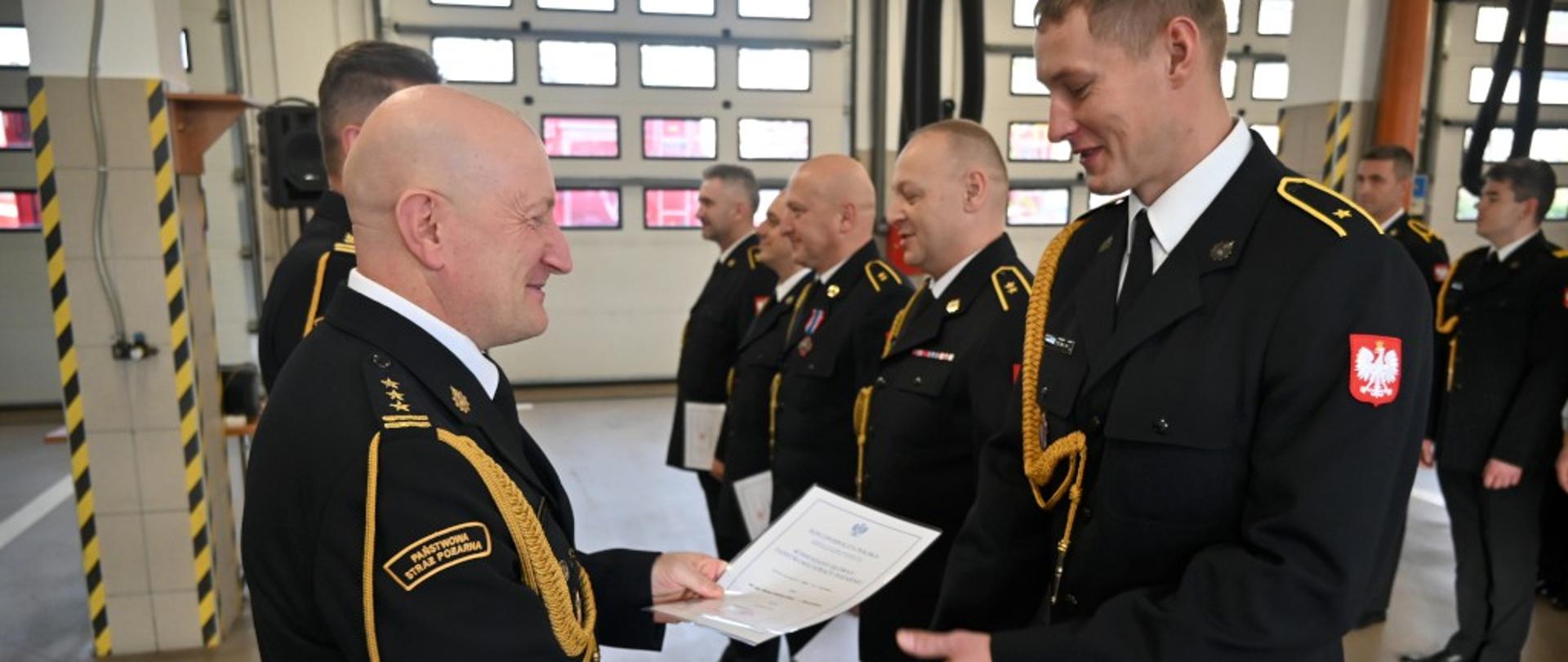 Z lewej Komendant Wojewódzki, z prawej w rzędzie wyróżnieni strażacy. Komendant składa gratulacje asp. Michałowi Erdmańskiemu z Bydgoszczy. Wszyscy ubrani w granatowe mundury wyjściowe bez nakryć głowy. Uroczystość odbywa się w garażach jednostki w Nakle.