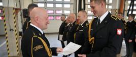 Z lewej Komendant Wojewódzki, z prawej w rzędzie wyróżnieni strażacy. Komendant składa gratulacje asp. Michałowi Erdmańskiemu z Bydgoszczy. Wszyscy ubrani w granatowe mundury wyjściowe bez nakryć głowy. Uroczystość odbywa się w garażach jednostki w Nakle.