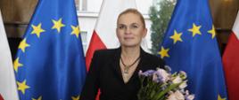 Minister Nowacka stoi na tle rzędu flag Polski i UE, w rękach trzyma bukiet kwiatów.