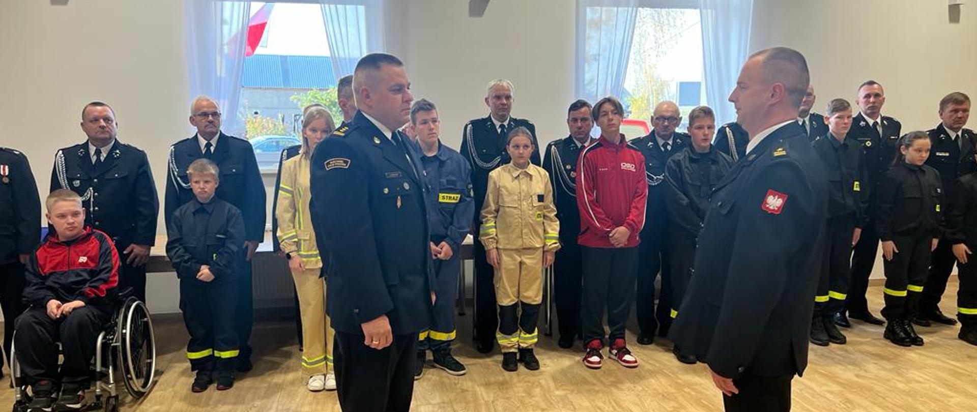 składanie meldunku strażakowi przez innego strażaka, w tle grupa strazaków i młodzieży w mundurach strażackich