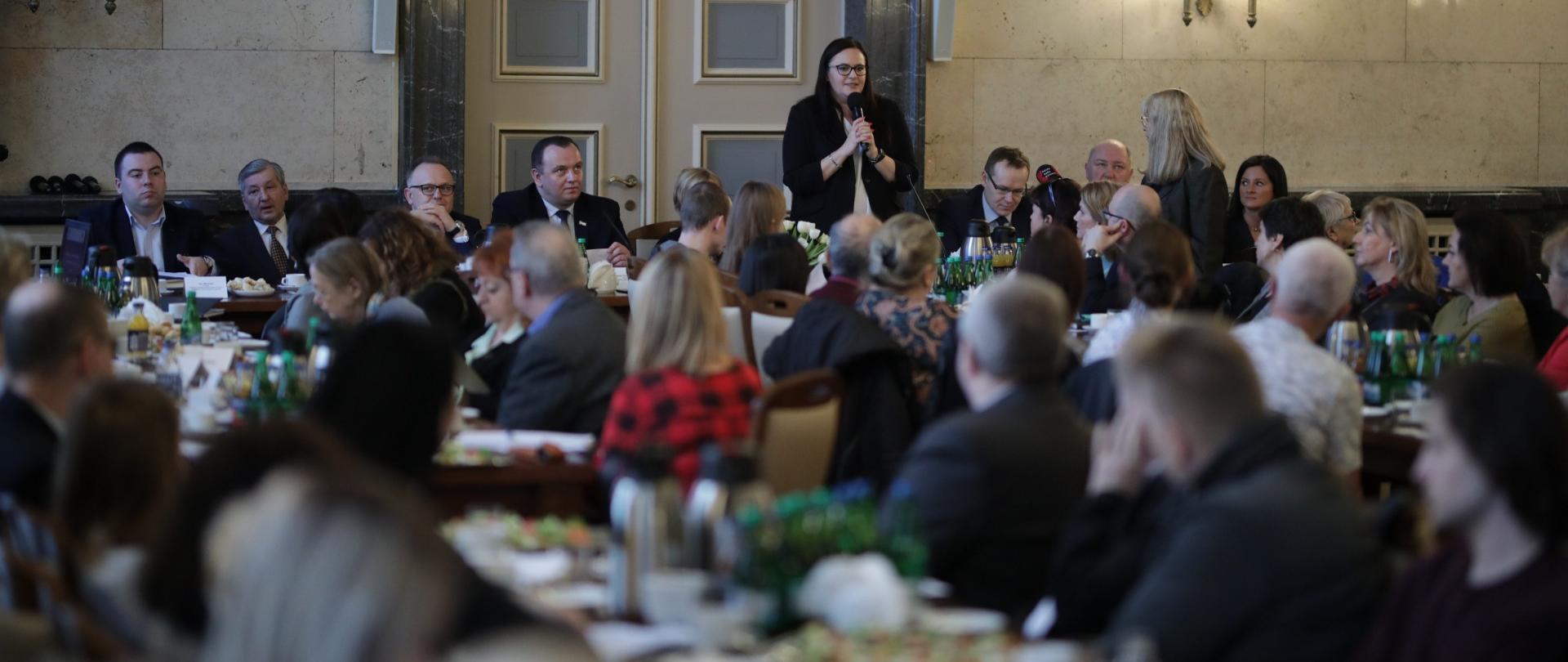 minister Małgorzata Jarosińska-Jedynak mówi do mikrofonu stojąc, pozostali uczestnicy spotkania siedzą wokół stołów