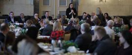 minister Małgorzata Jarosińska-Jedynak mówi do mikrofonu stojąc, pozostali uczestnicy spotkania siedzą wokół stołów