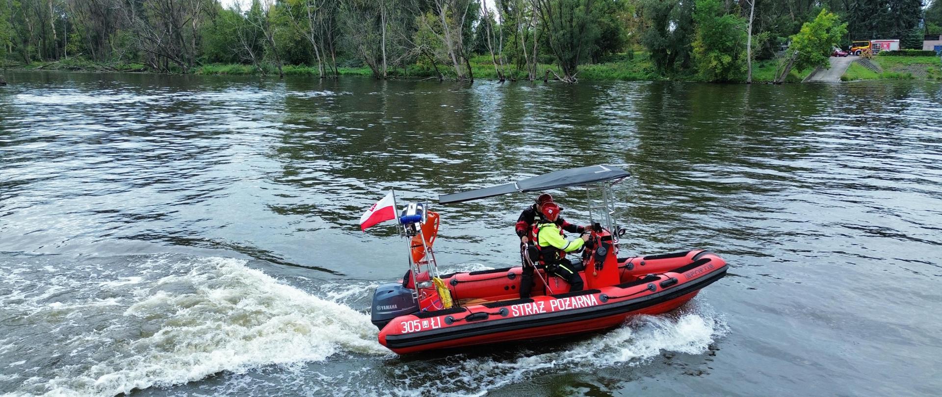 Strażacy znajdują się na łodzi motorowej płyną po rzece. Mają na sobie kamizelki ratunkowe. Na łodzi widnieje napis „STRAŻ POŻARNA”. W tle widać zalesiony brzeg rzeki.