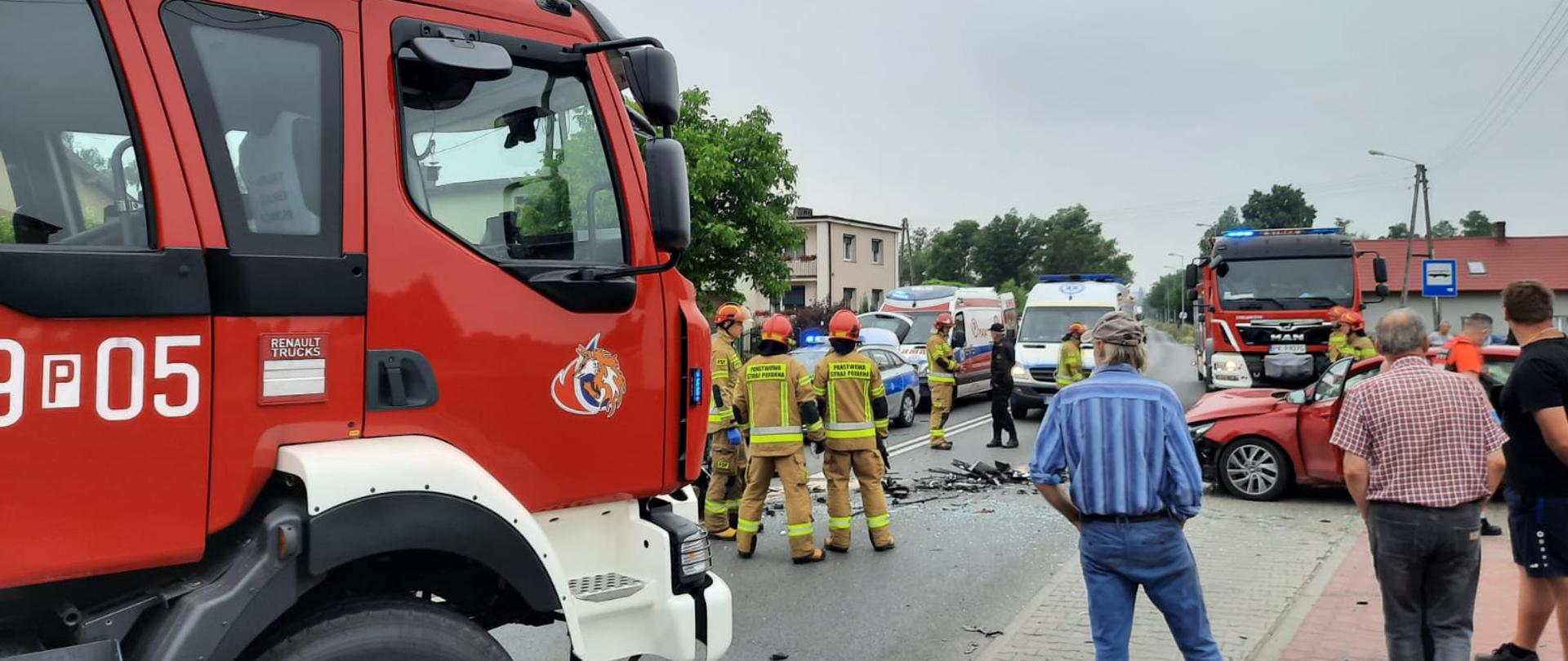 Na zdjęciu widać samochód gaśniczy OSP Kalisz-Lis. Pojazd jest w kolorze czerwonym, posiada sygnalizację ostrzegawczą. W centralnym miejscu rozbity pojazd, w kolorze czerwonym. Wokół ratownicy OSP i PSP ubrani w ubrania specjalne w kolorze pisakowym. Dalej karetki Państwowego Ratownictwa Medycznego. Niebo zachmurzone. Po prawej stronie osoby postronne. 