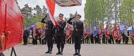 poczet sztandarowy z trzech strażaków ubranych w mundury wyjściowe osp umieszcza flagę państwową na koszu drabiny mechanicznej, w tle stoją poczty sztandarowe kilkunastu jednostek ochotniczych straży pożarnych