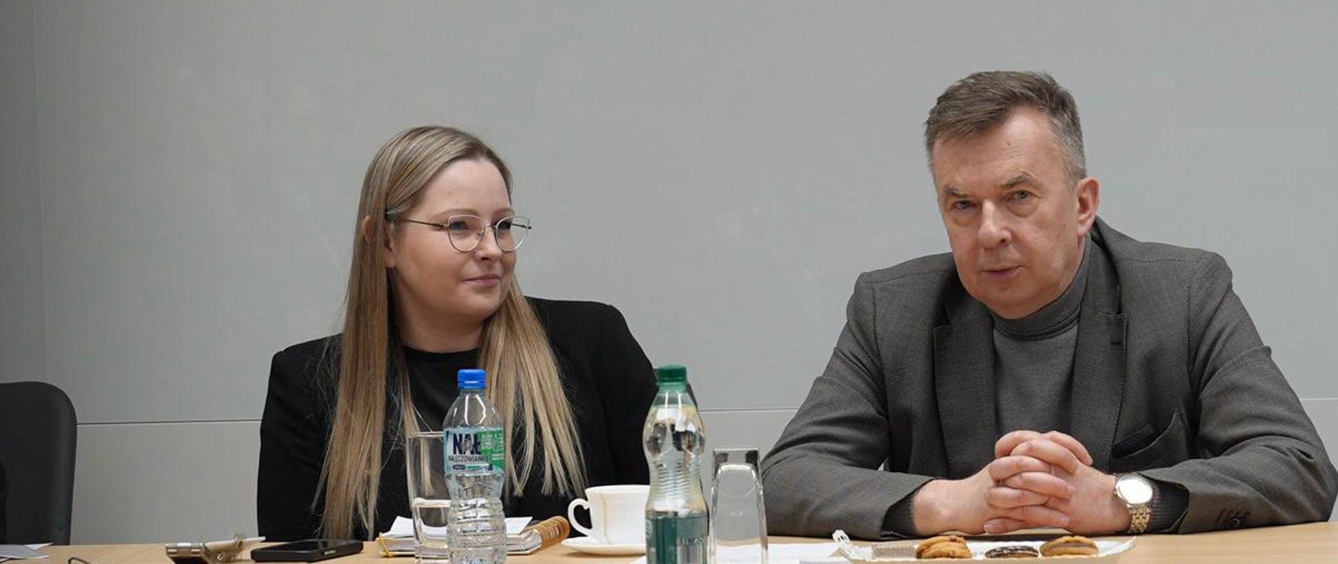 Minister Wieczorek siedzi za stołem i mówi, obok niego ubrana na czarno kobieta.