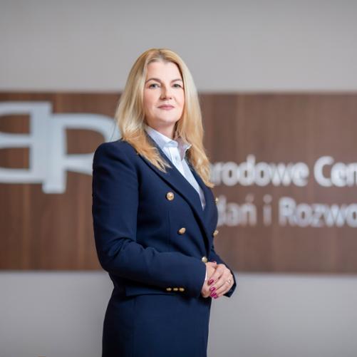 Na tle napisu Narodowe Centrum Badań i Rozwoju kobieta stojąca bokiem w granatowym żakiecie