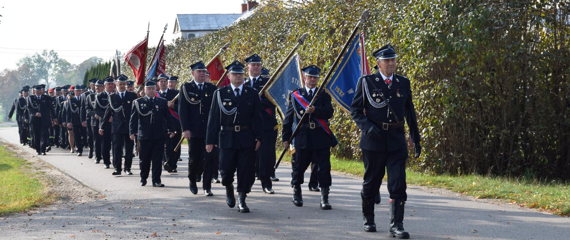Pododdział strażaków ubranych w umundurowanie galowe maszeruje ulicą. Na czele dowódca pododdziału, następnie osiem pocztów sztandarowych, za nimi pozostali strażacy. 
