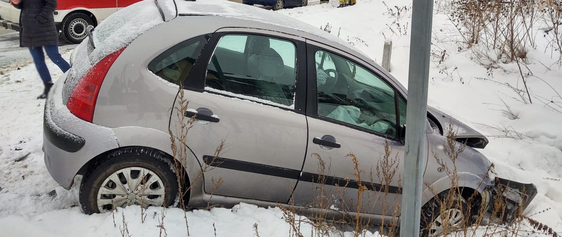 Samochód Citroen z rozbitym przodem poza drogą na śniegu. Obok stoi kobieta. W tle karetka oraz drugi samochód biorący udział w zdarzeniu. Obok drugiego samochodu stoją strażacy.