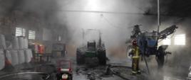 Widoczna zadymiona hala magazynowa, strażak pracujący w środku, spalony ciągnik rolniczy, widoczne inne maszyny rolnicze