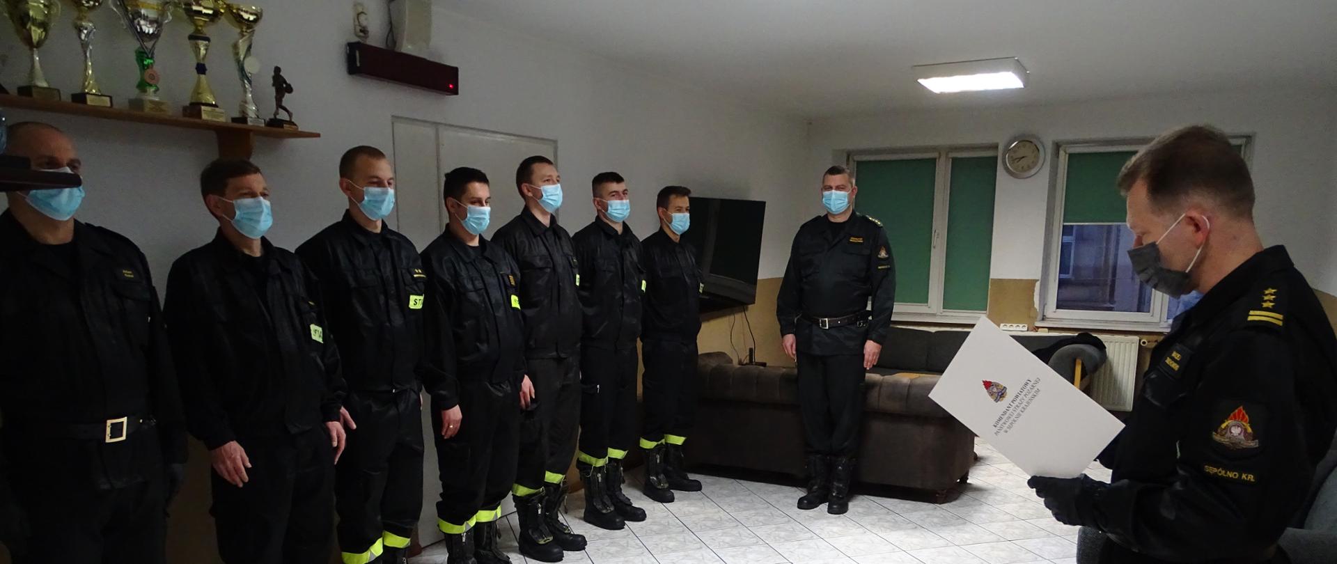 Zdjęcie wykonano w pomieszczeniu świetlicy budynku Komendy Powiatowej Państwowej Straży Pożarnej w Sępólnie Krajeńskim. Na zdjęciu Komendant Powiatowy starszy brygadier Maciej Suchomski w mundurze służbowym odczytuje rozkaz. Po lewej stronie zmiana ubrana w mundury koszarowe stoi na baczność. 