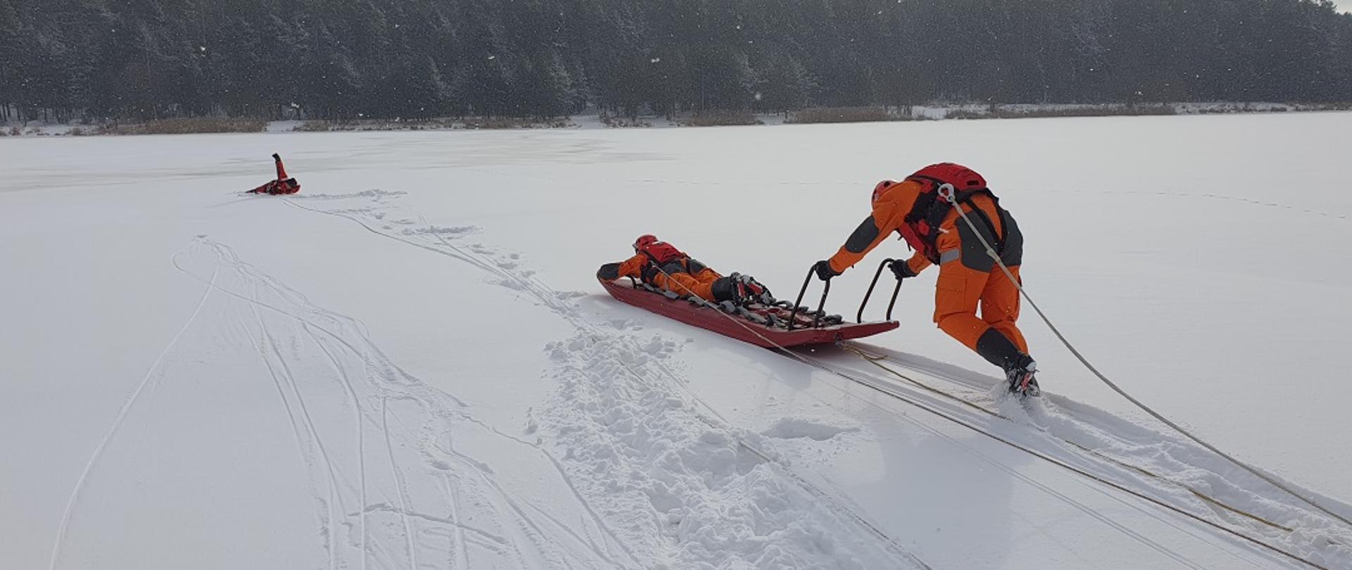 Na zdjęciu widzimy przybyłe na miejsce zdarzenie służby ratownicze. Dwóch ratowników Państwowej Straży Pożarnej ubranych w specjalne ubrania wypornościowe (które nie pozwolą im utonąć), zabezpieczeni linkami ratowniczymi przywiązanymi do pasów strażackich przy pomocy specjalnych sań lodowych udają się na pomoc osobie, pod którą załamał się lód. Do sań lodowych widzimy przywiązaną linkę. Dzięki niej, strażacy którzy zabezpieczają ratowników będą mogli ich szybko wyciągnąć na brzeg. 