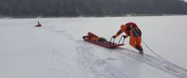 Na zdjęciu widzimy przybyłe na miejsce zdarzenie służby ratownicze. Dwóch ratowników Państwowej Straży Pożarnej ubranych w specjalne ubrania wypornościowe (które nie pozwolą im utonąć), zabezpieczeni linkami ratowniczymi przywiązanymi do pasów strażackich przy pomocy specjalnych sań lodowych udają się na pomoc osobie, pod którą załamał się lód. Do sań lodowych widzimy przywiązaną linkę. Dzięki niej, strażacy którzy zabezpieczają ratowników będą mogli ich szybko wyciągnąć na brzeg. 