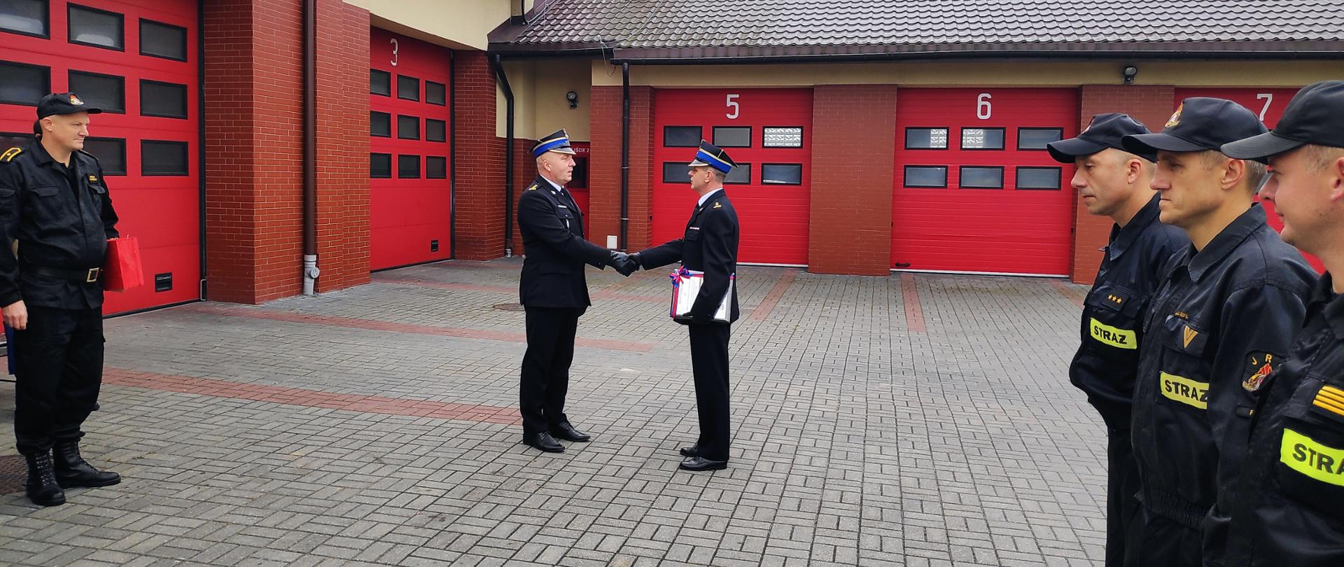 Zastępca Komendanta Powiatowego Państwowej Straży Pożarnej w Rypinie Pan brygadier Andrzej Górecki wręcza list gratulacyjny i składa życzenia Komendantowi Powiatowemu Państwowej Straż Pożarnej w Rypinie, Panu starszemu brygadierowi Grzegorzowi Łydkowskiemu. Za nimi czerwone wrota garażowe. Po prawej i lewej stronie na polbruku stoją strażacy w dwuszeregach. 