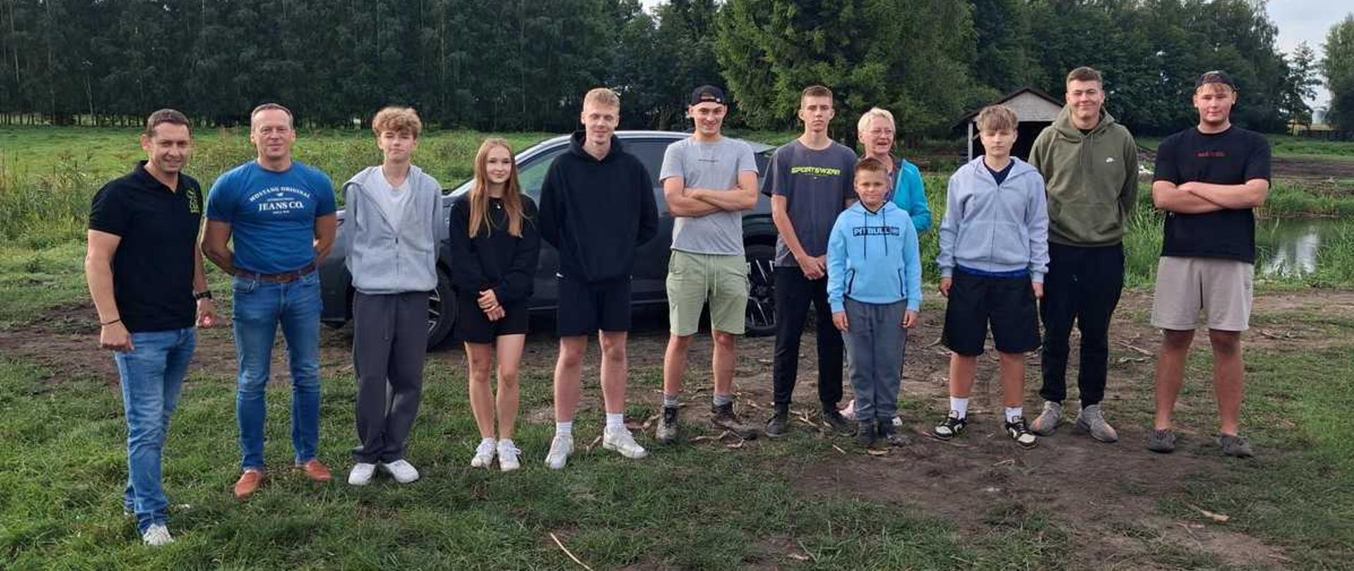 djęcie przedstawia grupę dwunastu osób stojących w jednym rzędzie na trawiasto-ziemniamym terenie pod gołym niebem. W tle widoczny jest ciemny samochód osobowy, ściana gęstego, zielonego lasu oraz fragment małego budynku po prawej stronie. Niebo jest zachmurzone, z jasnosarymi chmurami.Osoby na zdjęciu (od lewej do prawej)Mężczyzna w czarnej koszulce polo, niebieskich jeansach i białych sportowych butach. Stoi z uśmiechem, opierając lewą rękę na biodrze.Mężczyzna w niebieskiej koszulce z białym napisem „Mustang”, niebieskich jeansach i brązowych butach. Stoi z rękami opuszczonymi wzdłuż ciała.Młody chłopak w szarej rozpinanej bluzie z kapturem, białym t-shircie i ciemnosarych dresowych spodniach.Młoda dziewczyna z długimi, jasnymi włosami. Ma na sobie czarną bluzę, czarną spódniczkę lub spodenki i białe buty sportowe. Trzyma dłonie splecione przed sobą.Młody chłopak w czarnej bluzie z kapturem, ciemnych krótkich spodenkach i białych butach ze skarpetami.Młody chłopak w szarym t-shircie, jasnoniebieskich krótkich spodenkach i czapce z daszkiem założonej tył na przód. Stoi z ramionami skrzyżowanymi na piersi.Młody chłopak w ciemnogranatowej koszulce z napisami, ciemnych długich spodniach i czarnych butach.Mały chłopiec w jasnoniebieskiej bluzie z kapturem i ciemnych spodniach dresowych, stojący z przodu.Starsza kobieta w okularach i jaskrawoniebieskiej bluzie, stojąca tuż za małym chłopcem.Młody chłopak w jasnoszarej rozpinanej bluzie z kapturem, ciemnych krótkich spodenkach i sportowych butach.Młody chłopak w oliwkowej (zielonej) bluzie z kapturem, czarnych spodniach dresowych i ciemnych butach.Młody chłopak w czarnym t-shircie, szarych krótkich spodenkach i klapkach, stojący z ramionami skrzyżowanymi na piersi.Tło i otoczeniePierwszy plan: Nierówna, częściowo wydeptana ziemia z kępami zielonej trawy. Na ziemi leży długa, cienka gałąź lub sznurek.Drugi plan: Bezpośrednio za grupą stoi ciemnoszary samochód, zza którego wystają głowy niektórych osób. Po prawej stronie widać fragment małego, drewnianego lub murowanego domku z ciemnym dachem.Dalszy plan: Wysoka, zwarta linia zielonych drzew (las), która rozciąga się wzdłuż całego horyzontu.