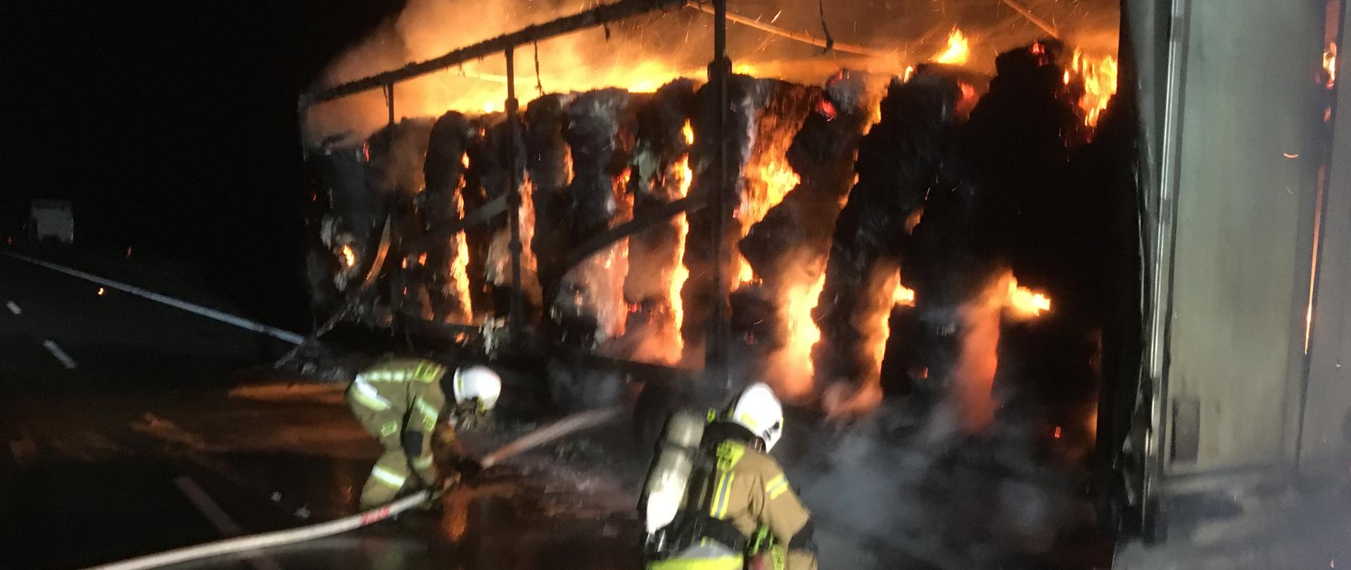 Naczepa samochodowa stoi na poboczu drogi w środku kostki papieru objęte ogniem - cała naczepa w płomieniach jeden strażak ubrany w sprzęt ODO trzyma w ręku wąż czerwony z którego leje die woda na źródło pożaru , obok drugi strażak z wężem 