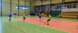 Zawodnicy rywalizujący podczas meczu futsalu w hali sportowej – drużyny w żółtych i niebieskich strojach są w trakcie dynamicznej akcji na boisku. Strażak z Kp PSP w Olecku w trakcie wykonywania podania.