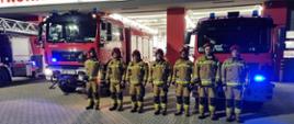 Zdjęcie przedstawia siedmiu strażaków stojących przed trzema wozami i strażnicą