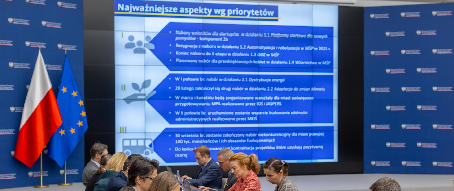 Grupa osób siedzi przy stole na dużej sali. 