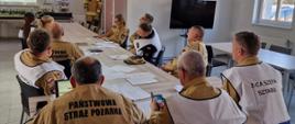 W sali szkoleniowej grupa strażaków Państwowej Straży Pożarnej siedzi przy dużym stole i uczestniczy w odprawie. Na stole znajdują się dokumenty, laptopy i telefony. Na ścianie wyświetlana jest prezentacja, a obok wiszą herby. W tle stoi manekin w stroju strażackim oraz telewizor.