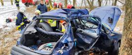 na pierwszym planie samochód osobowy Peugot 206 w rowie po wypadku z drzewem zderzenie boczne od strony pasażera, za samochodem strażacy pochyleni zbierają sprzęt po akcji, tle samochód pożarniczy stojący na drodze