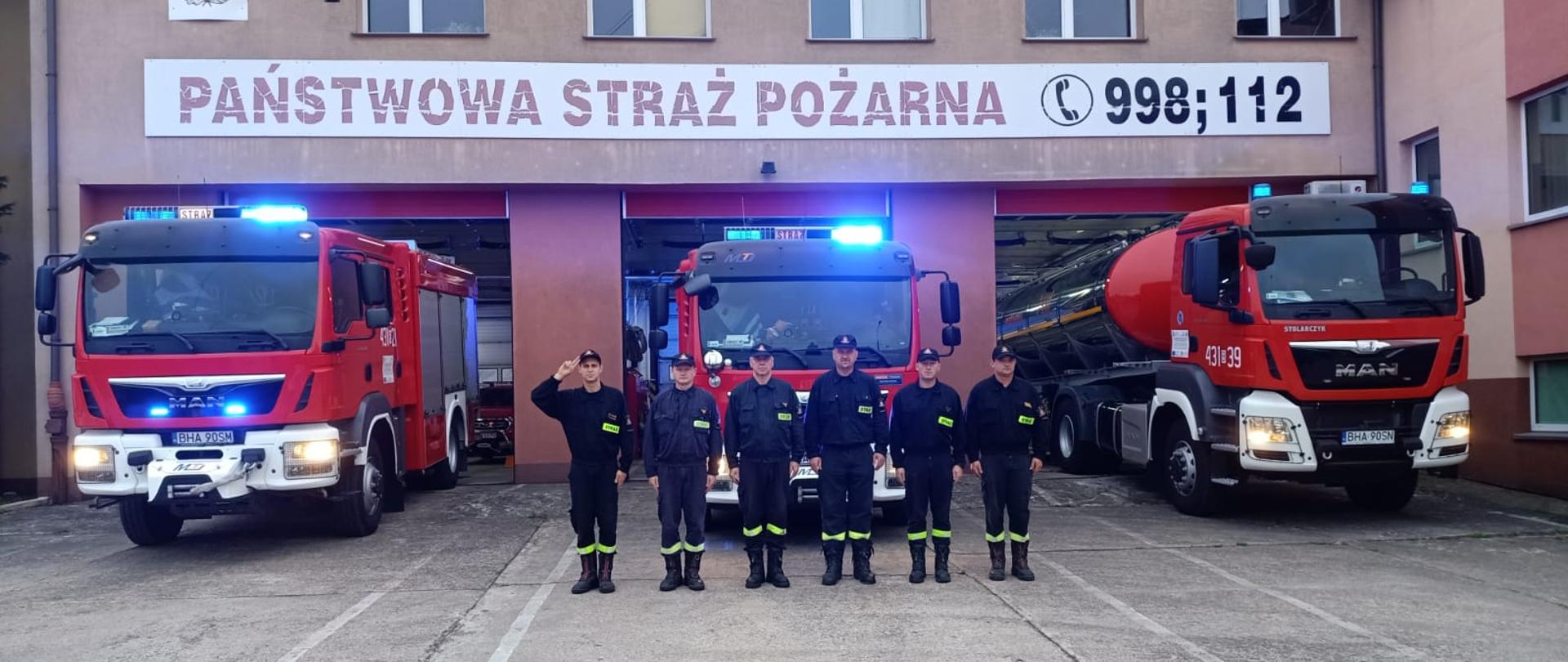 Obok czerwonego strażackiego wozu bojowego stoi kilku strażaków w czarnych mundurach.