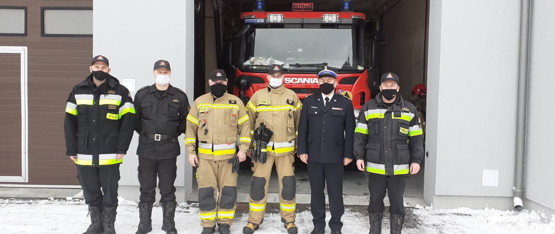 6 funkcjonariuszy PSP stoi w rzędzie przed garażami Posterunku JRG w Różanie. Jeden garaż jest otwarty, jest tam zaparkowany samochód gaśniczy Scania. 3 funkcjonariuszy jest w ubraniach KDS, 2 w ubraniach specjalnych, 1 - Mazowiecki Komendant Wojewódzki w ubraniu wyjściowym.