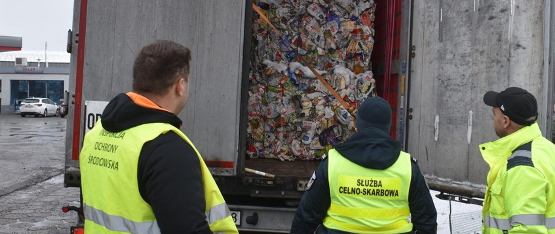 Trzech funkcjonariuszy w odblaskowych kamizelkach, w tym jeden ze służby celno-skarbowej i jeden z inspekcji ochrony środowiska, kontroluje otwartą naczepę ciężarówki. W środku znajdują się sprasowane odpady, głównie plastikowe opakowania.