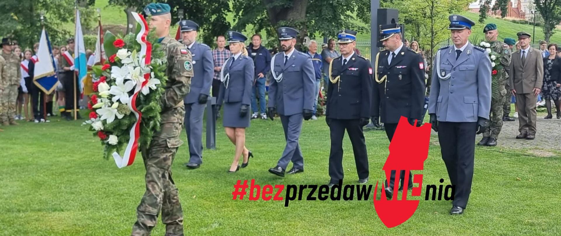 na pierwszym planie żołnierz z wieńcem z kwiatów biało-czerwonych. Za nim delegacja służb mundurowych - 4 policjantów i dwóch strażaków. W tle goście wydarzenia, poczty sztandarowe.