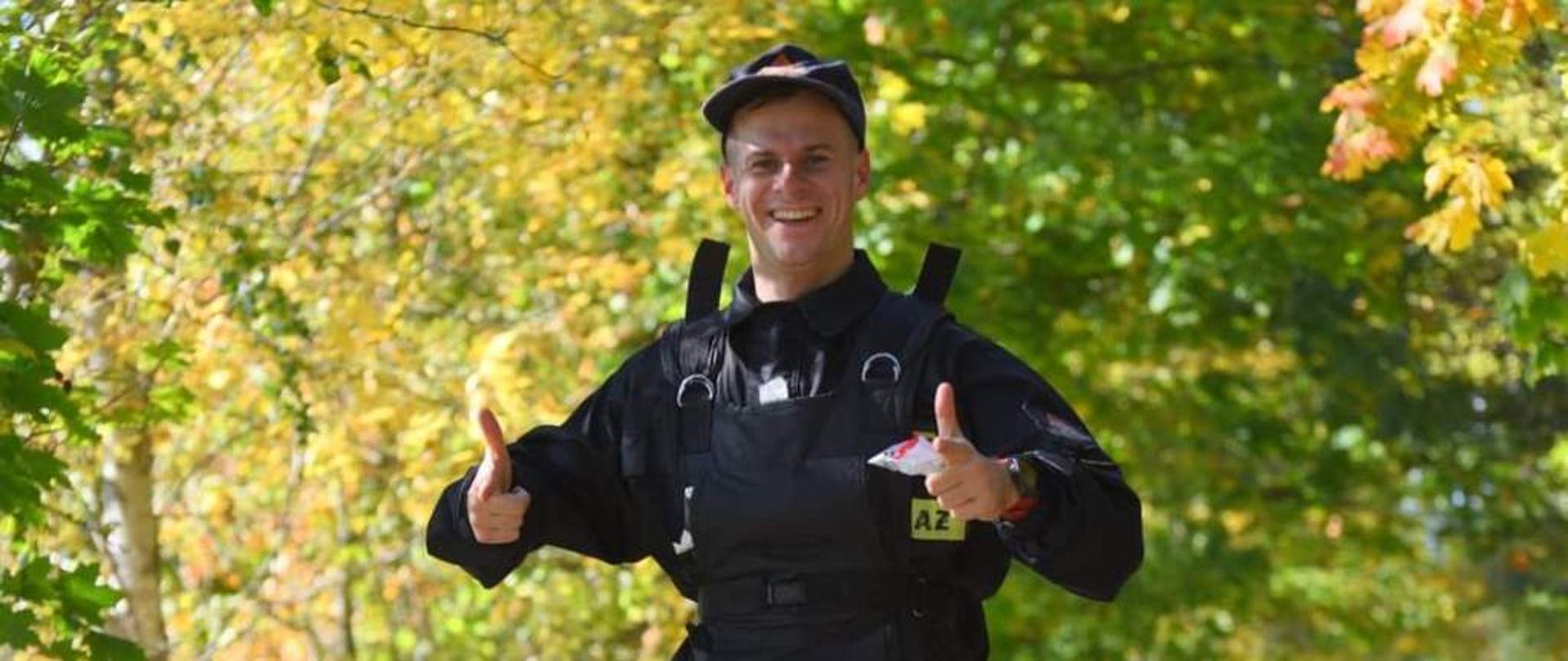 Na pierwszym planie uczestnik biegu militarnego na trasie biegu, sekcyjny Wojciech Plebanek, ubrany w ubranie koszarowe strażaka, na sobie ma również kamizelkę obciążającą 5 kilogramów. Trasa przebiega drogą leśną, w oddali inni uczestnicy biegu.