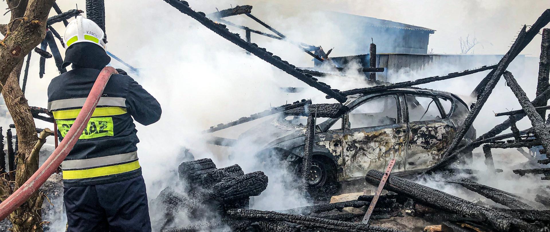 Na zdjęciu znajduje się spalony samochód oraz elementy drewnianej szopy. Po lewej stronie stoi strażak, który trzyma wąż strażacki i dogasza pogorzelisko.