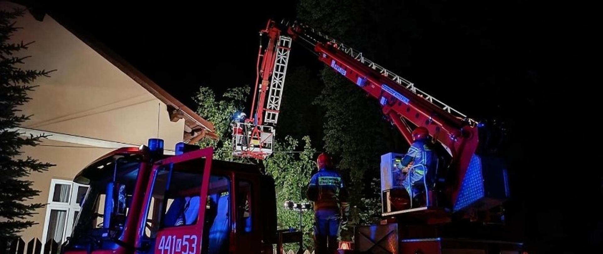 Na zdjęciu widoczny na pierwszym planie podnośnik hydrauliczny a w nim strażacy pomagający w doraźnym zabezpieczeniu uszkodzonego dachu na budynku poprzez zakładanie plandeki.