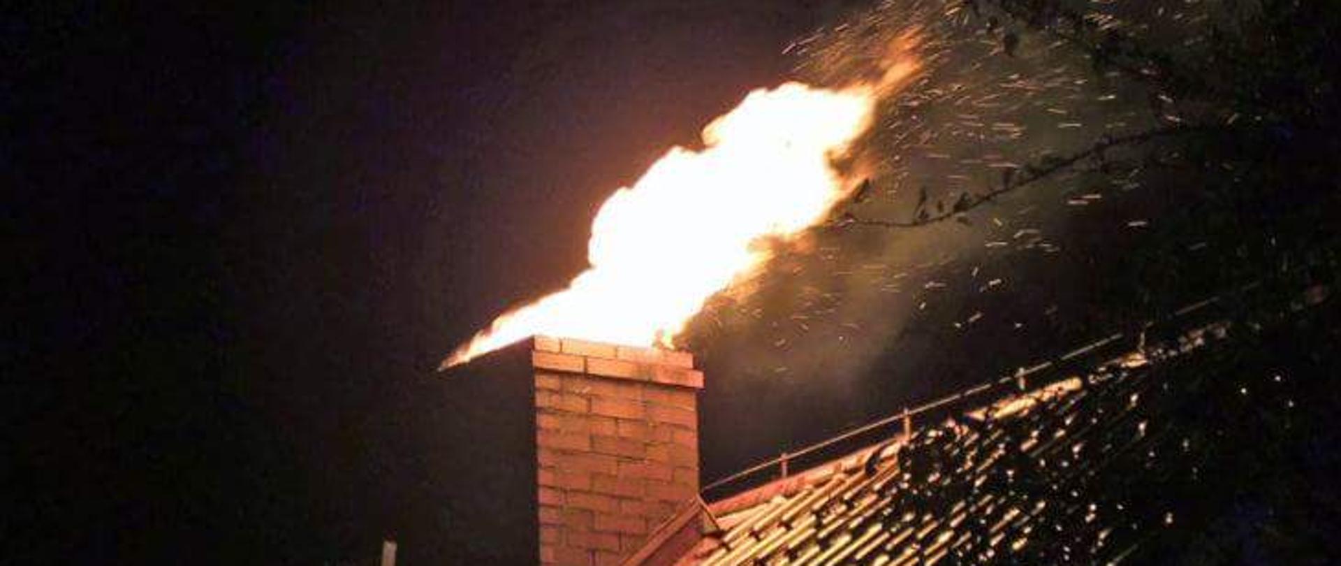 Zdjęcie przedstawia pożar sadzy w kominie. Na zdjęciu widać dach budynku i komin z którego wydobywa się ogień. Pora nocna. 