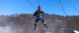 Strażacy ćwiczą ewakuację z wyciągu krzesełkowego. Na pierwszym planie strażak zabezpieczony w uprząż ewakuacyjną typu "trójkąt" zostaje spuszczany bezpiecznie na linie na teren gruntu.