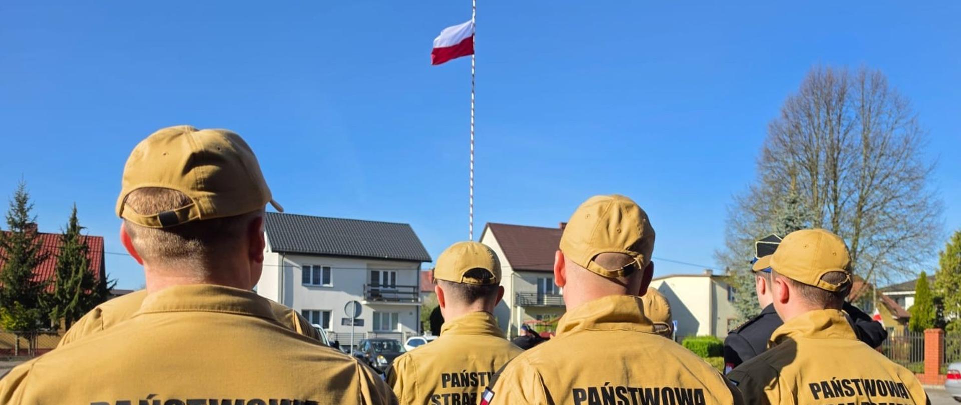 Grupa strażaków w piaskowych mundurach służbowych stoi tyłem, patrząc w stronę polskiej flagi na wysokim maszcie. Na plecach ich bluz widnieje czarny napis „PAŃSTWOWA STRAŻ POŻARNA”, a w tle widać domy jednorodzinne i bezchmurne, niebieskie niebo. Uroczystość z okazji Dnia Flagi Rzeczypospolitej.