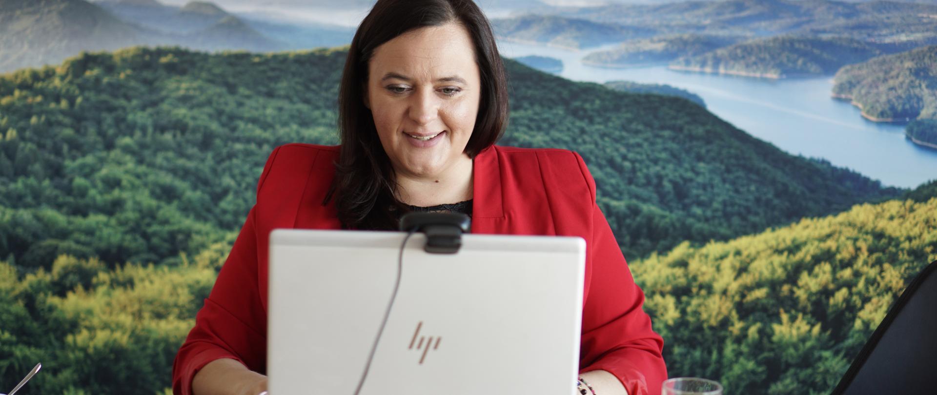 Minister Jarosińska-Jedynak przed monitorem komputera, w tle krajobraz las i rzeka