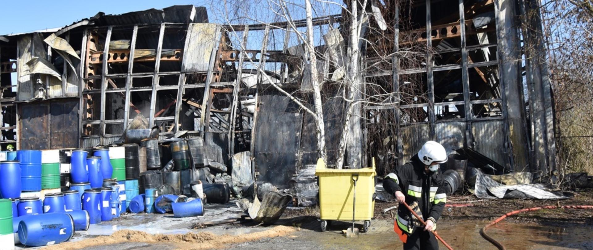 Funkcjonariusz Straży Pożarnej prowadzi prace porządkowe na utwardzonym terenie. W tle spalona, dwupiętrowa hala magazynowa oraz beczki 200 l, które nie uległy spaleniu.