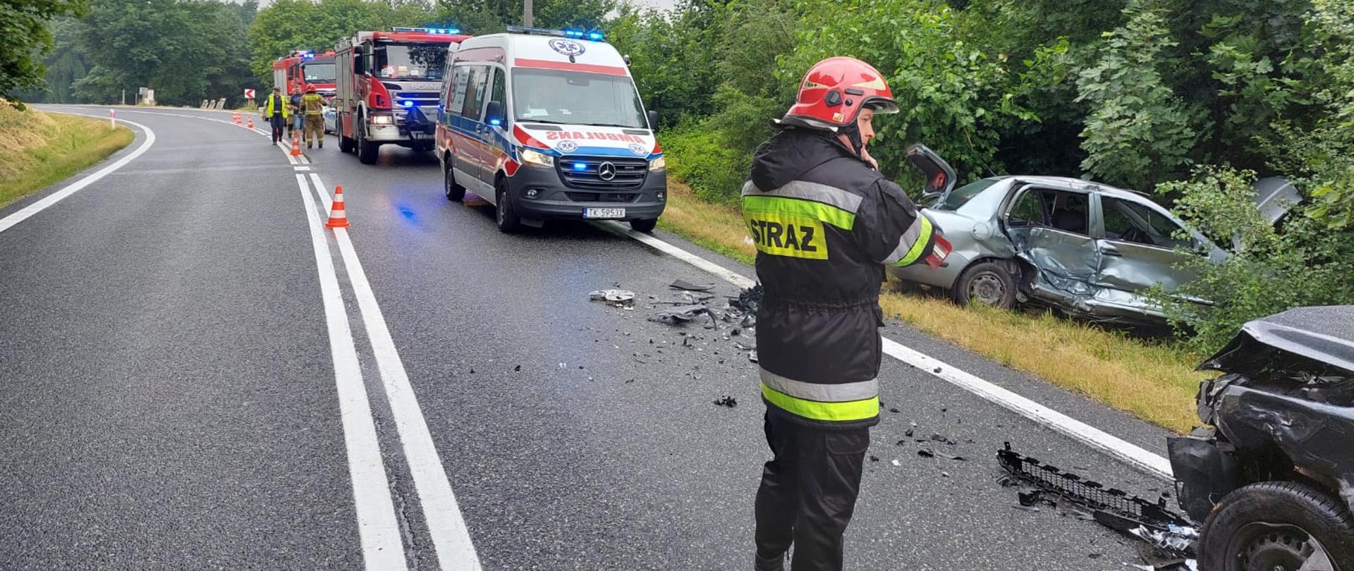 Zdjęcie po prawej stronie przedstawia dwa rozbite samochody osobowe. Jeden z nich znajduję się w rowie, drugi na drodze krajowej na jednym pasie. Po lewej stronie widać strażaka w umundurowaniu dowódczo-sztabowym oraz karetkę pogotowia i dwa samochody ratowniczo-gaśnicze z włączoną sygnalizacją alarmową świetlną. 