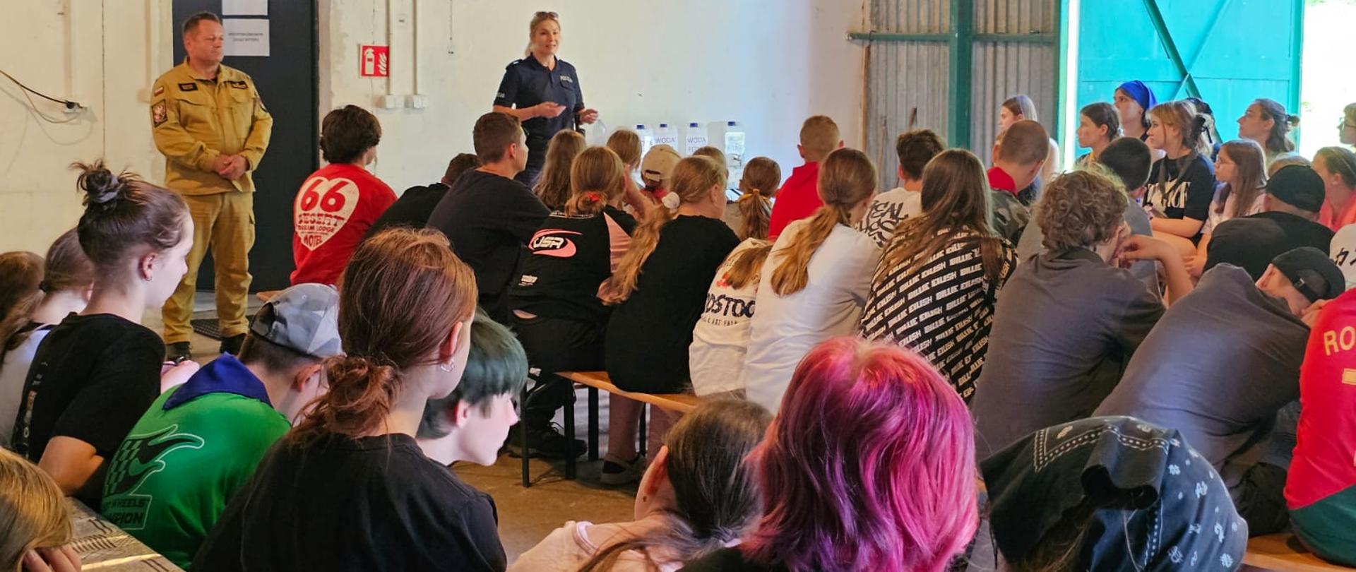 Zdjęcie przedstawia uczestników obozu harcerskiego pod namiotami oraz kadrę opiekunów podczas pogadanki edukacyjnej dla dzieci, młodzieży i osób dorosłych prowadzonej przez funkcjonariusza Komendy Powiatowej Państwowej Straży Pożarnej w Gołdapi oraz przedstawiciela Komendy Powiatowej Policji w Gołdapi