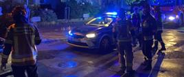 ratownicy PSP stoją na skrzyżowaniu, w tle radiowóz Policji oraz wóz pożarniczy, oba na sygnałach błyskowych.