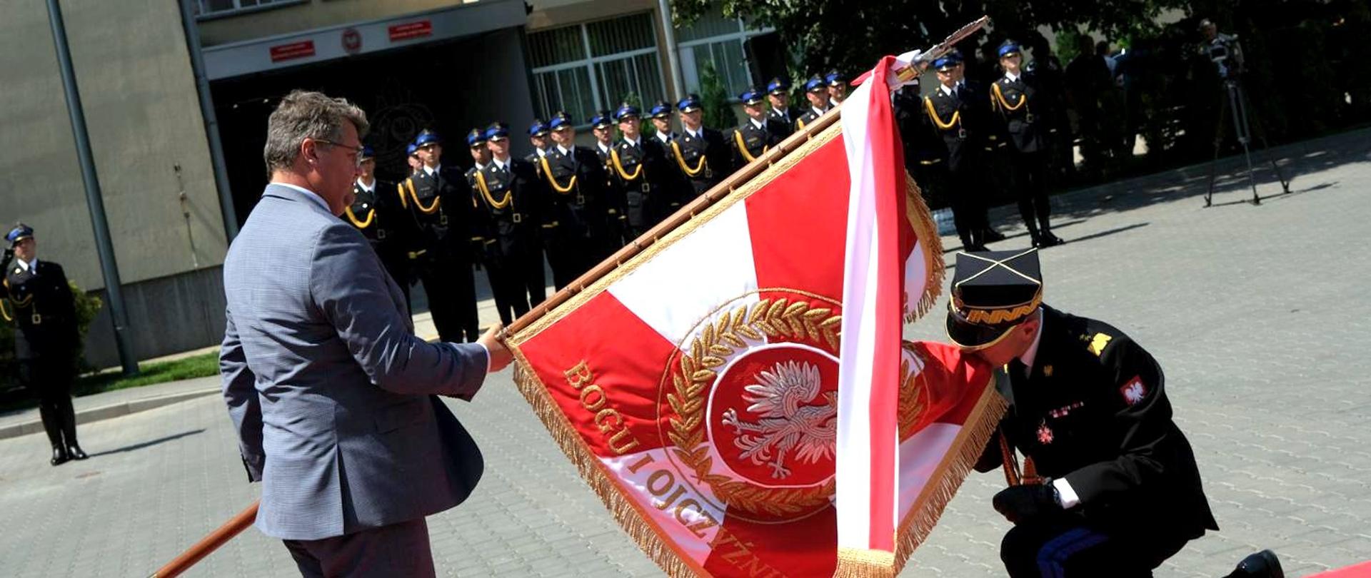 Wiceminister spraw wewn. i admin. trzyma sztandar pochylony do przodu, przed sztandarem klęka komendant główny PSP całując róg sztandaru