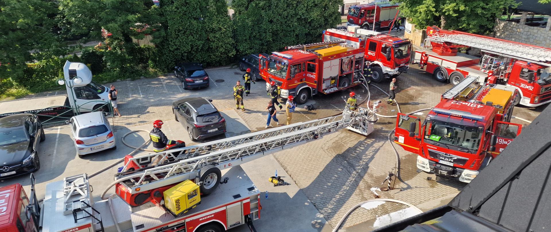 Widok z dachu na parking hotelowy. Na parkingu widać 6 czerwonych wozów bojowych straży pożarnej, rozwinięte węże pożarnicze, strażaków w ubraniach bojowych. 