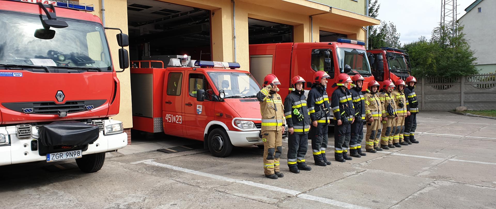 Zdjęcie przedstawia stojących w szeregu strażaków. Z tyłu za nimi cztery wozy strażackie.