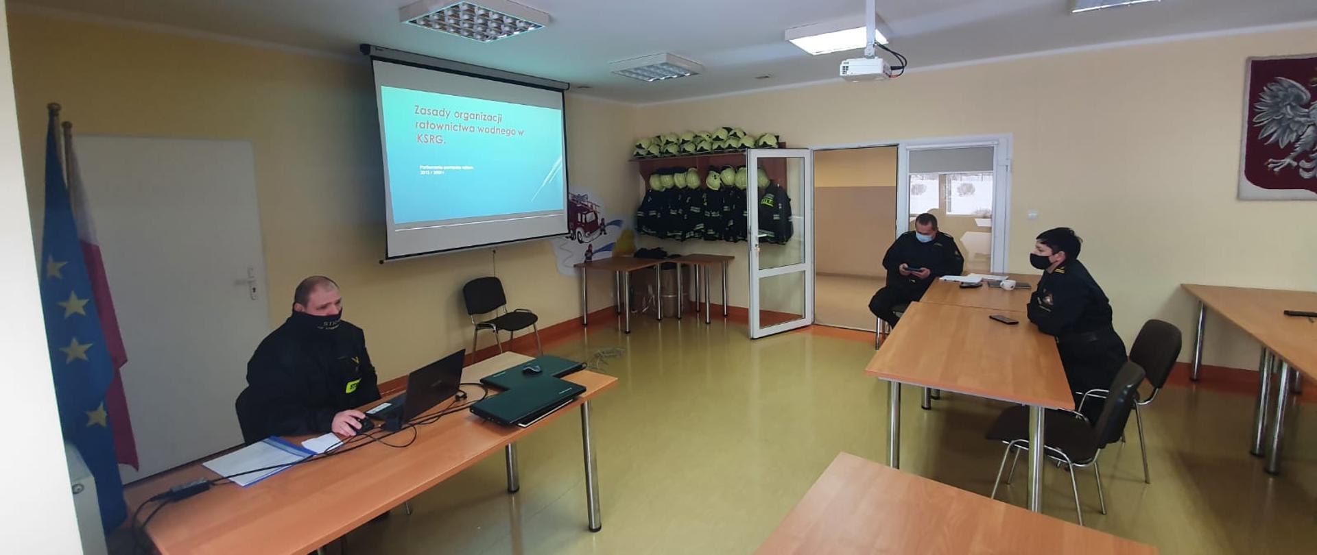 Funkcjonariusze uczestniczący w szkoleniu, widoczny obraz prezentacji na tablicy multimedialnej umiejscowionej po lewej stronie kadru. Prowadzący siedzi na wprost słuchaczy z zachowaniem dystansu z uwagi na pandemię. 