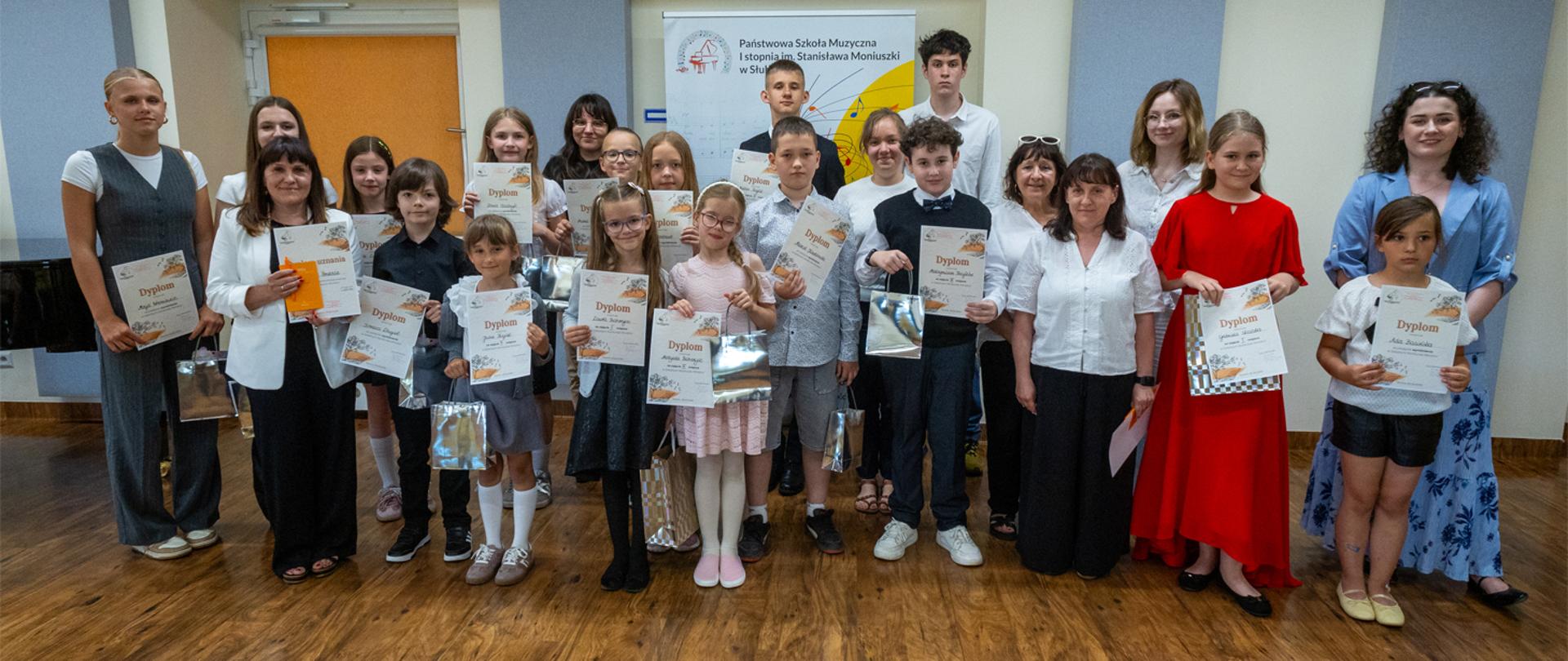 Kilkunastoosobowa grupa nauczycieli i uczniów, stoją w auli szkoły, uczniowie trzymają w rękach dyplomy.