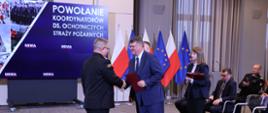 Strażak w mundurze wyjściowym odbiera gratulacje od sekretarza stanu w Ministerstwie Spraw Wewnętrznych i Administracji obok stoi komendant główny Państwowej Straży Pożarnej za nimi stoi kobieta z teczkami. 