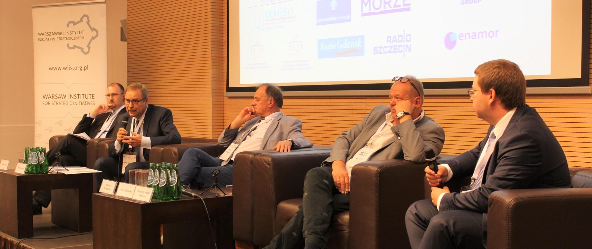 Na podium, w fotelach siedzi czterech prelegentów oraz prowadzący. Jeden z nich przemawia.