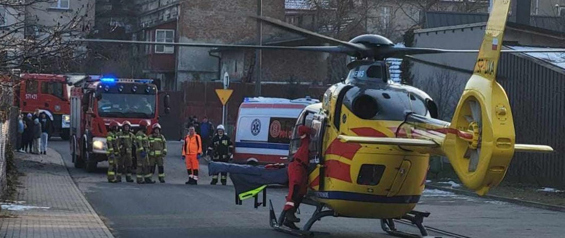 Ulica Wodna w Jutrosinie. Na jezdni stoi śmigłowiec Lotniczego Pogotowia Ratunkowego. Z jego lewej strony znajduje się członek załogi. Przed śmigłowcem zaparkowana jest karetka pogotowia oraz dwa wozy strażackie. Pomiędzy pojazdami znajdują się strażacy i ratownik medyczny. W tle kamienice.